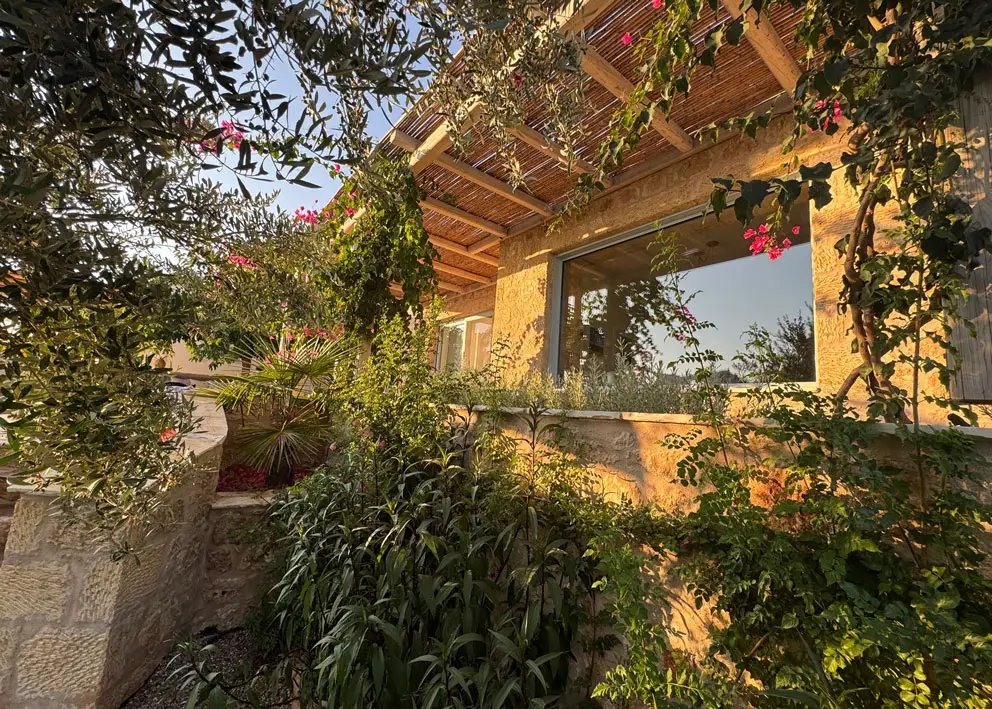 Außenansicht eines Ferienhauses in Griechenland, umgeben von üppiger Vegetation und Blumen, typisch für die griechische Landschaft. Das Ferienhaus ist mit einem Strohdach versehen.